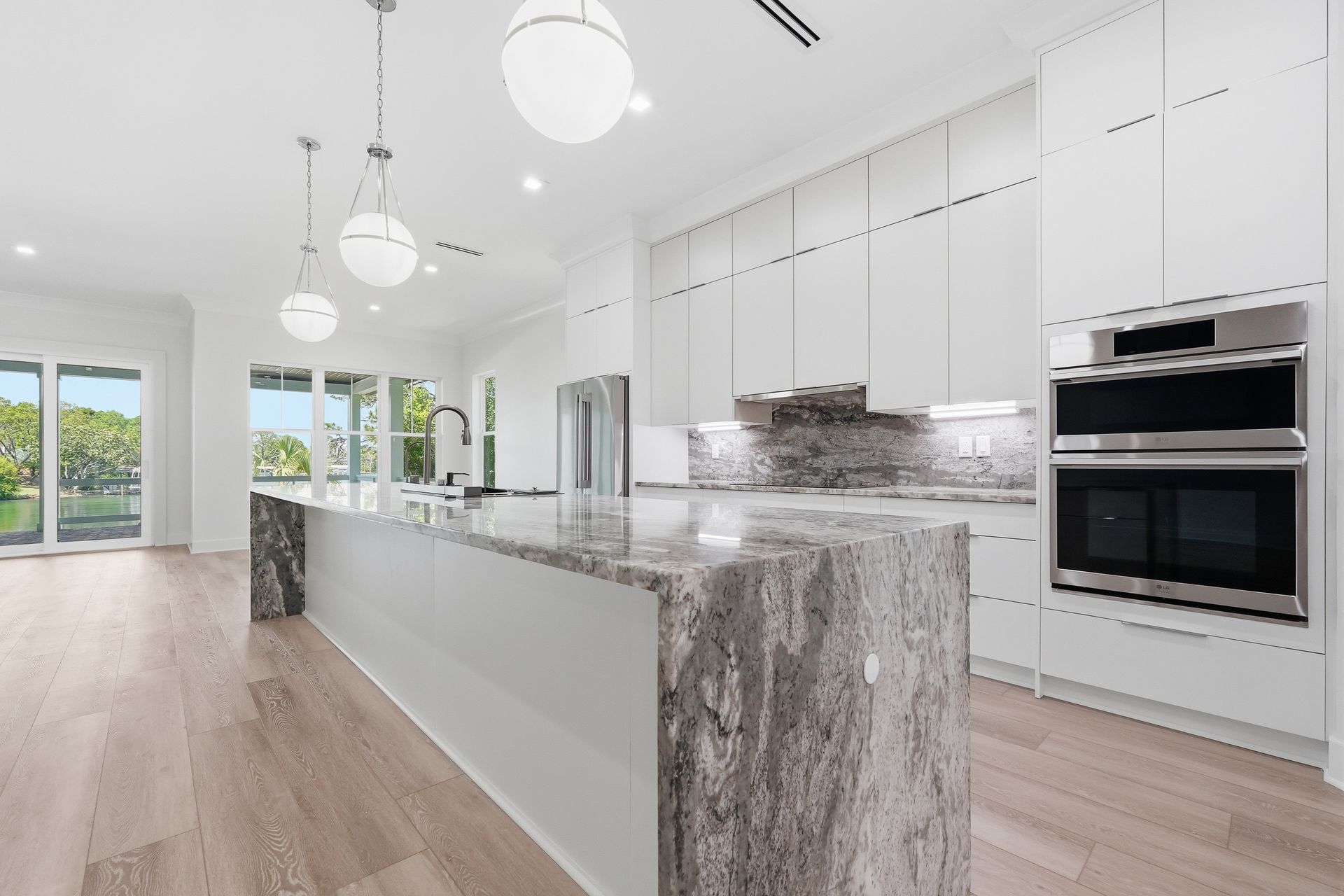 Open kitchen in a new waterfront home with a marble waterfall island, white cabinetry and water visible through the far windows